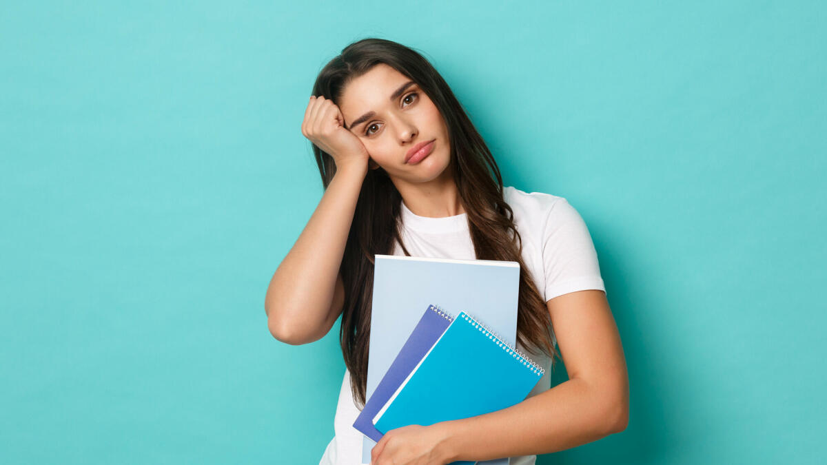 Porträt einer müden, hübschen Studentin, die Studienmaterial und Notizbücher in der Hand hält und erschöpft aussieht, stehend vor blauem Hintergrund © Mix and Match Studio / 500px / Getty Images